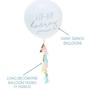 K&ouml;nnte beinhalten: Wei&szlig;er Ballon mit 91 cm Durchmesser mit dem aufgedruckten Text "HIP-HIP hooray". Der Ballon hat eine lange dekorative Quaste mit 9 Quasten in Rosa, Blau, Gelb und Gold.