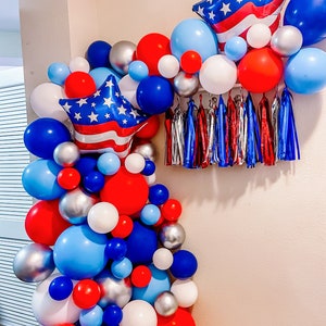 K&ouml;nnte beinhalten: Ein rot-wei&szlig;-blauer Ballonbogen mit sternf&ouml;rmigen Ballons und Luftschlangen. Der Bogen besteht aus Ballons in verschiedenen Gr&ouml;&szlig;en und Farben.