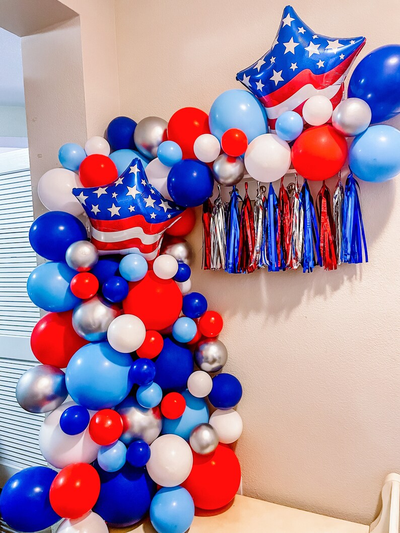 K&ouml;nnte beinhalten: Ein rot-wei&szlig;-blauer Ballonbogen mit sternf&ouml;rmigen Ballons und Luftschlangen. Der Bogen besteht aus Ballons in verschiedenen Gr&ouml;&szlig;en und Farben.
