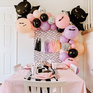 Op de afbeelding: Een roze tafel met een zwart-wit gestreept tafelkleed is gedekt voor een Halloween-feest. De tafel is versierd met zwarte en paarse ballonnen, zwarte vleermuizen en een zwart-wit pompoen-middelpunt.