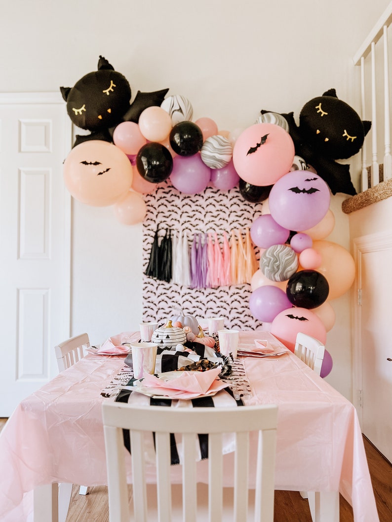 Op de afbeelding: Een roze tafel met een zwart-wit gestreept tafelkleed is gedekt voor een Halloween-feest. De tafel is versierd met zwarte en paarse ballonnen, zwarte vleermuizen en een zwart-wit pompoen-middelpunt.