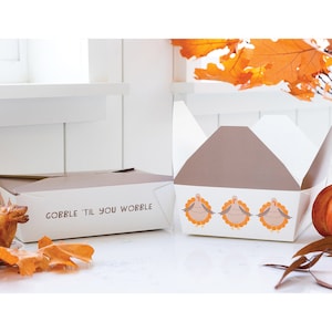 以下が含まれることがあります： 白と茶色の感謝祭をテーマにした食品ボックス。1つのボックスは「GOBBLE 'TIL YOU WOBBLE」のテキストで閉じられています。もう1つのボックスは開いており、3羽の漫画の七面鳥が表示されています。背景には紅葉とカボチャがあります。