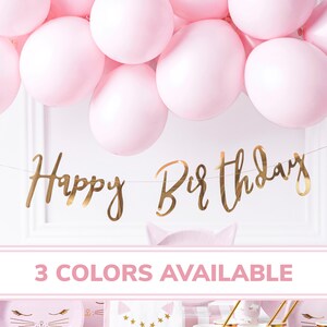 May include: Gold foil Happy Birthday banner with pink balloons in the background. The banner is strung on a clear string. The text "3 COLORS AVAILABLE" is at the bottom of the image.