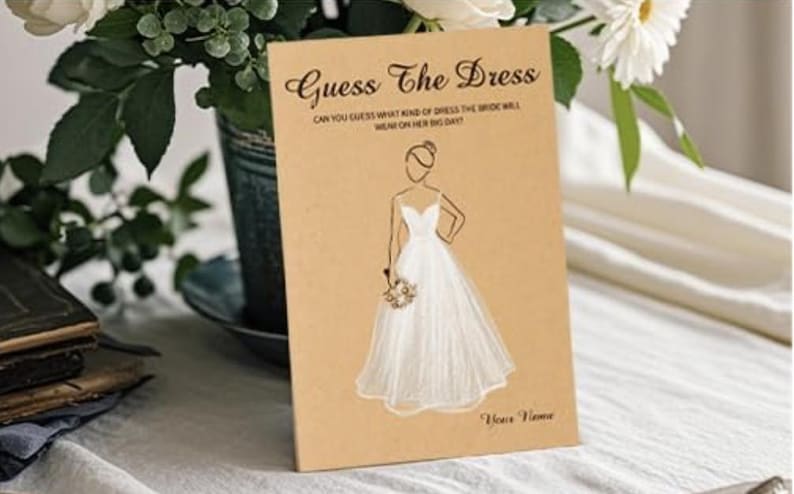 May include: A rectangular tan card featuring the black text Guess The Dress and Can you guess what kind of dress the bride will wear on her big day? with a central line drawing of a person in a white gown holding a bouquet and the text Your Name.