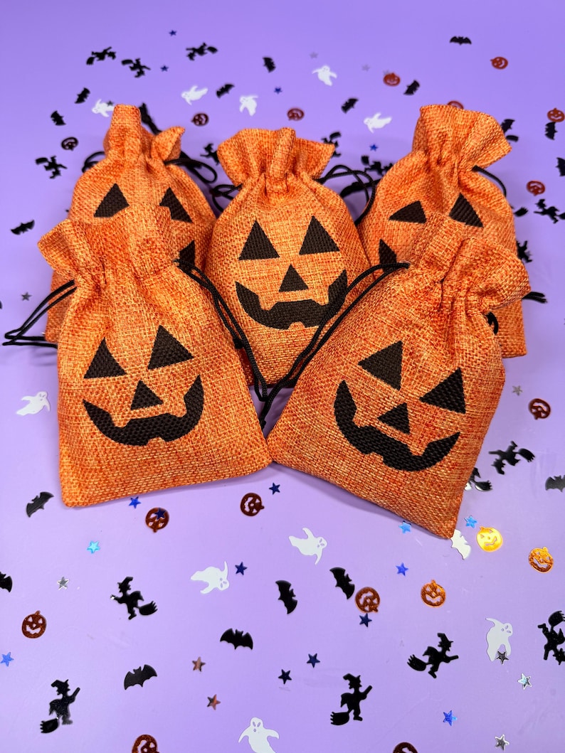 May include: A set of five orange burlap drawstring bags, each featuring a black jack-o'-lantern face print. The bags have a textured fabric weave and black cord closures at the top, arranged in a cluster on a light purple surface with confetti.