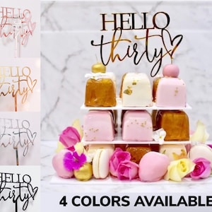 以下が含まれることがあります： 「Hello Thirty」というテキストが書かれたケーキトッパーで、黒い筆記体でハートが付けられています。トッパーは、ローズゴールド、ゴールド、シルバー、ブラックの4色展開です。ケーキトッパーは、マカロン、花、小さなケーキで飾られた3段のケーキが置かれた白いケーキスタンドの上にあります。