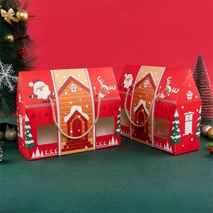 May include: Two red Christmas-themed gift boxes shaped like gingerbread houses. Each box features a Santa Claus and reindeer illustration, a window, and a rope handle. The boxes are decorated with festive details like trees and snow.