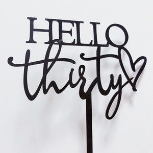 以下が含まれることがあります： 黒いアクリル製のケーキトッパーで、「Hello Thirty」というテキストが筆記体で書かれており、最後にハートが付いています。トッパーは黒い棒に付けられています。