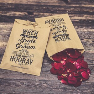 May include: Two brown paper bags with the text "WHEN THE Bride and Groom WALK YOUR WAY. TOSS THIS IN THE AIR AND SHOUT HOORAY" and "WHEN THE Bride and Groom HOORAY" printed on them. One bag is filled with red rose petals.