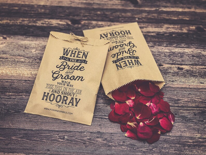 May include: Two brown paper bags with the text "WHEN THE Bride and Groom WALK YOUR WAY. TOSS THIS IN THE AIR AND SHOUT HOORAY" and "WHEN THE Bride and Groom HOORAY" printed on them. One bag is filled with red rose petals.