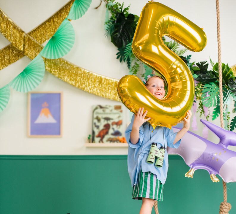 Pu&ograve; includere: Un bambino tiene un grande palloncino dorato a forma di numero cinque davanti al viso. Il palloncino &egrave; parzialmente gonfiato e ha una finitura lucida. Il bambino indossa una camicia blu e pantaloncini verdi. Sullo sfondo ci sono piante verdi e decorazioni.