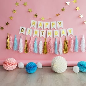 Pode incluir: Cena de festa de anivers&aacute;rio com fundo rosa. S&atilde;o exibidas guirlandas de estrelas douradas e uma faixa com a frase "HAPPY BIRTHDAY". Tassels coloridos e decora&ccedil;&otilde;es em favo de mel, juntamente com bal&otilde;es brancos, completam o ambiente festivo.