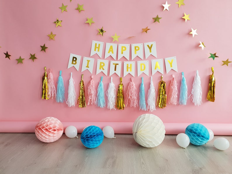 Pode incluir: Cena de festa de anivers&aacute;rio com fundo rosa. S&atilde;o exibidas guirlandas de estrelas douradas e uma faixa com a frase "HAPPY BIRTHDAY". Tassels coloridos e decora&ccedil;&otilde;es em favo de mel, juntamente com bal&otilde;es brancos, completam o ambiente festivo.