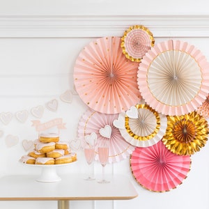 Puede incluir: Abanicos de papel rosa y dorado sobre un fondo blanco. Un soporte para pastel con donas decoradas con glaseado rosa y las palabras "She Said Yes" en la parte superior.