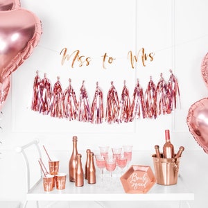 May include: Rose gold bridal shower decorations with a banner that says "Miss to Mrs." and a rose gold ice bucket with bottles of wine. There are also rose gold balloons, cups, and a sign that says "Bride Squad".