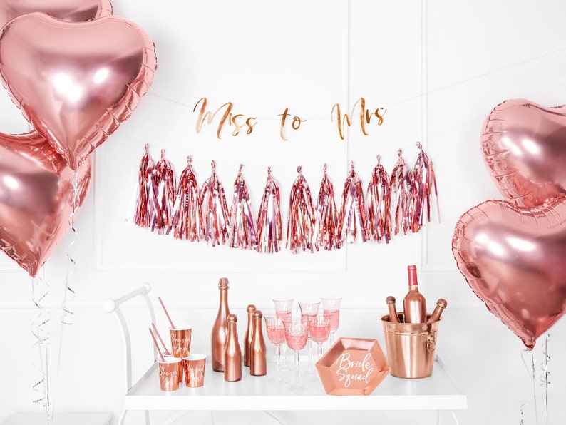 May include: Rose gold bridal shower decorations with a banner that says "Miss to Mrs." and a rose gold ice bucket with bottles of wine. There are also rose gold balloons, cups, and a sign that says "Bride Squad".
