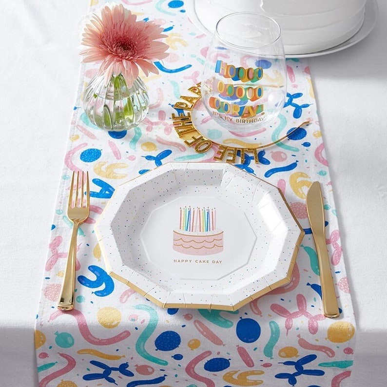 May include: A white paper party plate with a pink cake with rainbow candles and the text "Happy Cake Day". The plate is on a white table runner with a colorful balloon pattern. There is a gold fork and knife on the table.