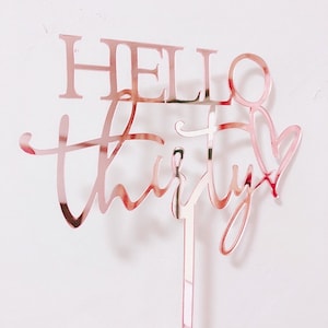 以下が含まれることがあります： 「Hello Thirty」と書いた、ローズゴールドのアクリル製のケーキトッパー。数字の「Thirty」は「Hello」よりも大きなフォントサイズです。