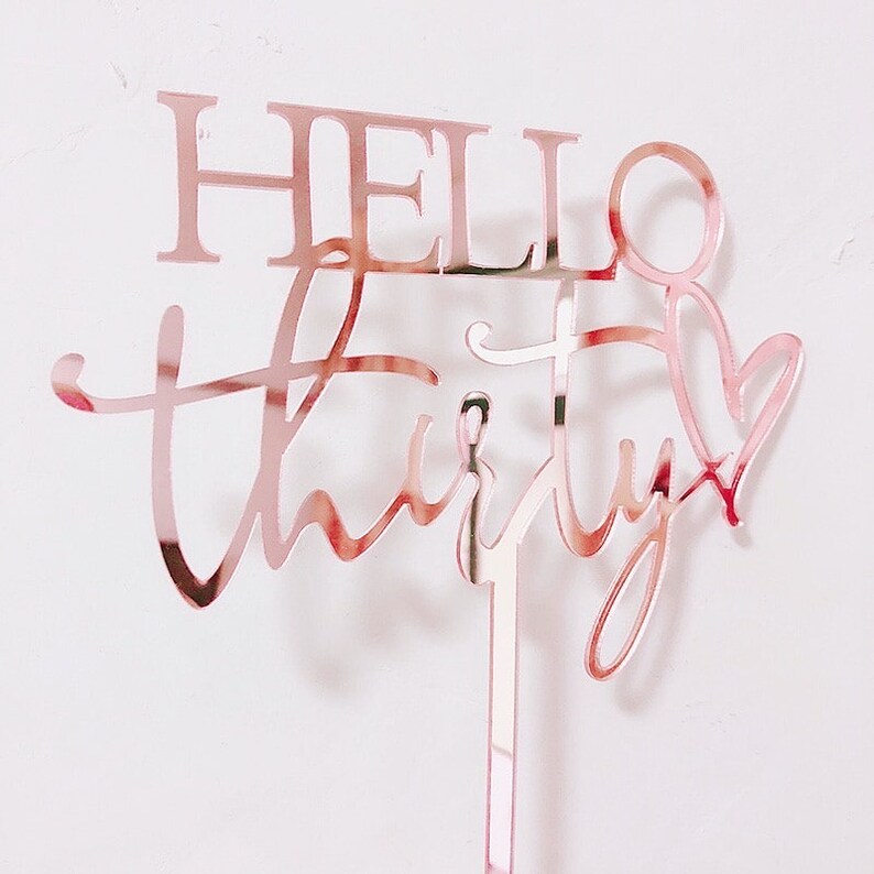 以下が含まれることがあります： 「Hello Thirty」と書いた、ローズゴールドのアクリル製のケーキトッパー。数字の「Thirty」は「Hello」よりも大きなフォントサイズです。