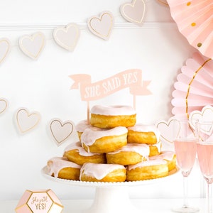 Puede incluir: Una pila de donuts cubiertos con glaseado rosa y una pancarta que dice "She Said Yes!" Los donuts est&aacute;n en un soporte para pasteles blanco. Hay decoraciones en forma de coraz&oacute;n rosa y dorado y abanicos de papel rosa en el fondo.