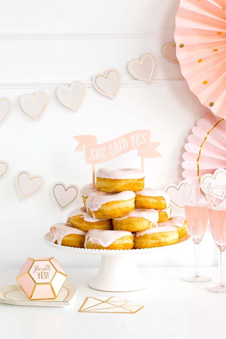 Puede incluir: Una pila de donuts cubiertos con glaseado rosa y una pancarta que dice "She Said Yes!" Los donuts est&aacute;n en un soporte para pasteles blanco. Hay decoraciones en forma de coraz&oacute;n rosa y dorado y abanicos de papel rosa en el fondo.