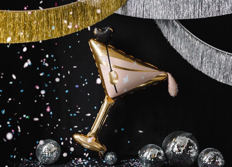Pu&ograve; includere: Un palloncino di lamina dorata e rosa a forma di bicchiere da martini con coriandoli che cadono intorno. Il palloncino &egrave; decorato con stelle bianche e un nastro rosa. Sullo sfondo ci sono sfere disco argentate.