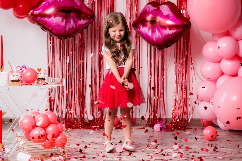 Op de afbeelding: Een jong meisje in een rode rok en een wit shirt staat voor een rode en witte achtergrond met roze en rode ballonnen. Confetti valt om haar heen.