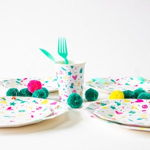 May include: A festive party setting with white plates, a cup, and pom-poms. The plates and cup feature a colorful pattern of stars, hearts, and holiday icons. Green and pink pom-poms are scattered around the plates. Green plastic utensils are in the cup.