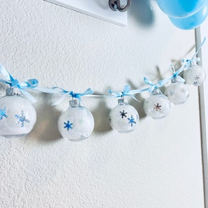 May include: A garland of clear, round ornaments with snowflake designs and blue ribbons. The ornaments are strung together with a light blue ribbon and hang against a white wall. Blue and purple balloons are visible in the background.
