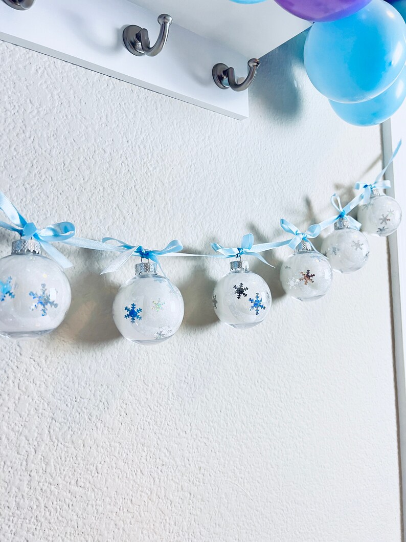 May include: A garland of clear, round ornaments with snowflake designs and blue ribbons. The ornaments are strung together with a light blue ribbon and hang against a white wall. Blue and purple balloons are visible in the background.
