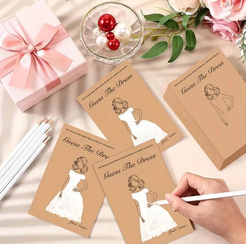 May include: A set of tan Guess The Dress game cards featuring a line art illustration of a person in a white dress, accompanied by a pink gift box with a satin ribbon, a glass bowl filled with red and white pearls, and white pencils.