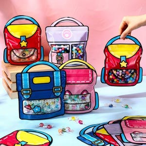 May include: Colorful, cartoon-style backpack-shaped bags filled with candies. The bags are red, pink, blue, and purple with yellow accents and black outlines. Some bags have a star or flower design. A hand holds one of the bags.