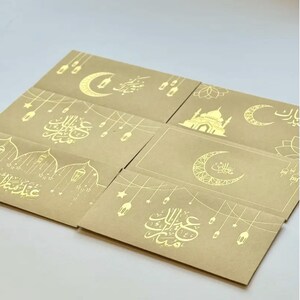 May include: A collection of six beige greeting cards with gold foil designs. The cards feature crescent moons, mosque illustrations, and Arabic calligraphy. The designs include hanging lanterns and stars, creating a festive and celebratory theme.
