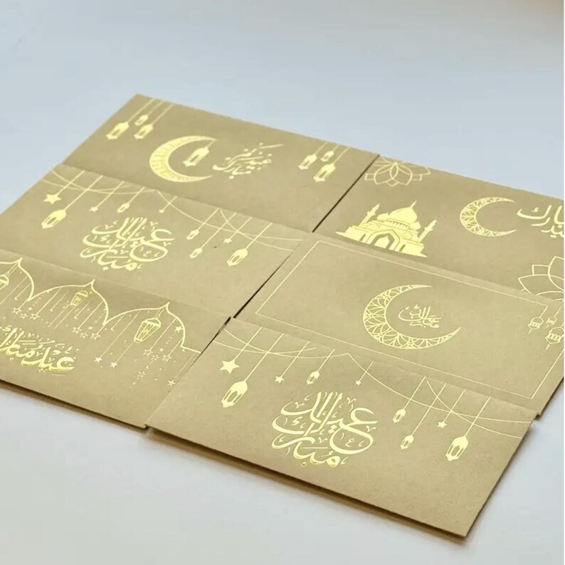 May include: A collection of six beige greeting cards with gold foil designs. The cards feature crescent moons, mosque illustrations, and Arabic calligraphy. The designs include hanging lanterns and stars, creating a festive and celebratory theme.