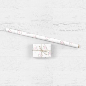 May include: A white gift box wrapped in white paper with a repeating pattern of pink Christmas lights and snowflakes. The box is tied with twine and is sitting on a white wooden surface.
