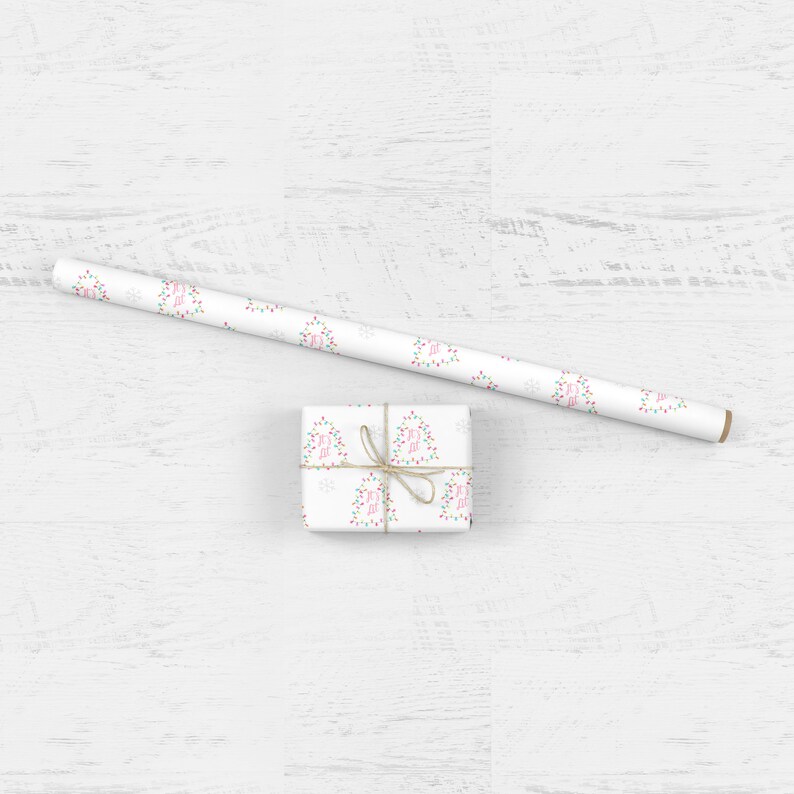May include: A white gift box wrapped in white paper with a repeating pattern of pink Christmas lights and snowflakes. The box is tied with twine and is sitting on a white wooden surface.