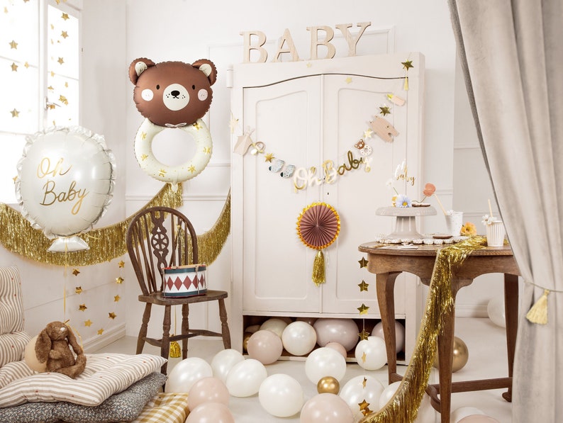 May include: A baby shower party decoration set with a brown bear balloon, a white balloon with the text "Oh Baby", gold fringe, and a wooden sign that says "BABY".
