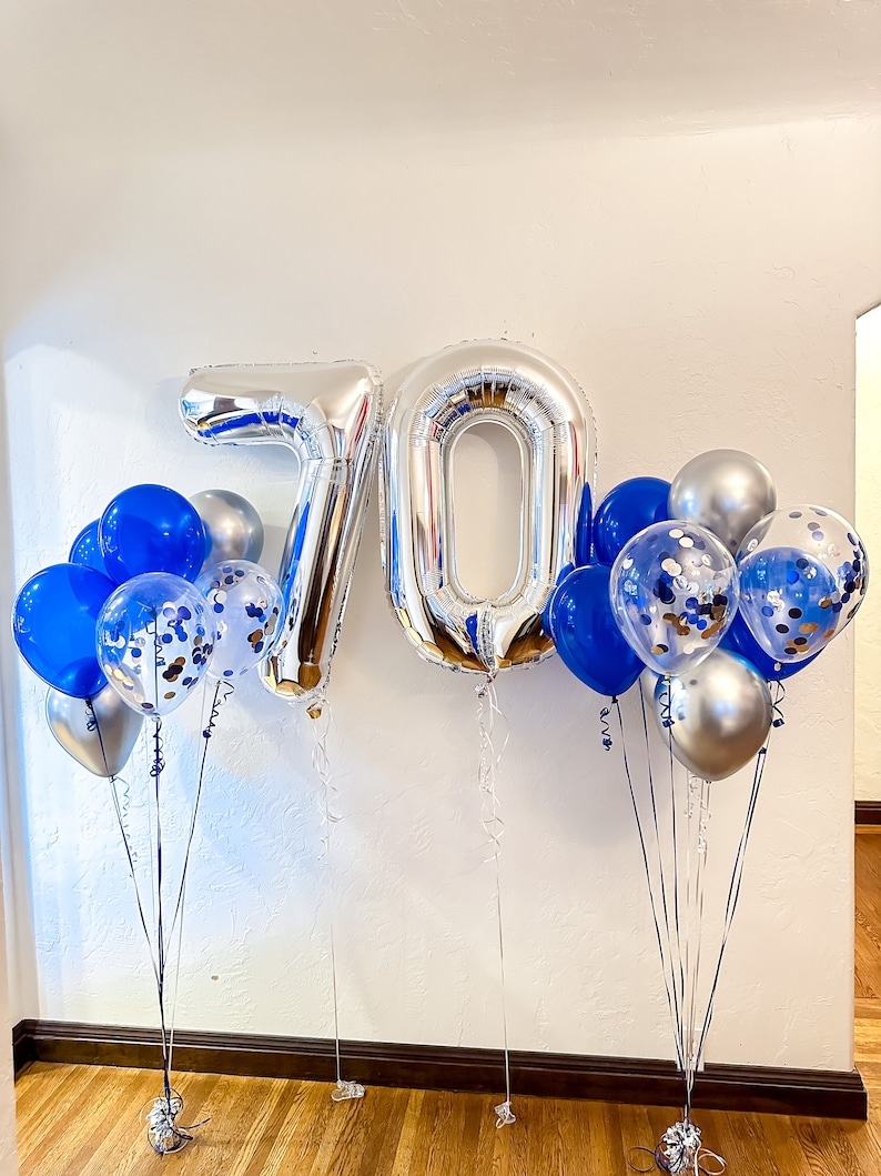 Może przedstawiać: Srebrne i niebieskie balony z konfetti, tworzące liczbę 70. Balony są związane ze sobą przezroczystą sznurkiem i przymocowane do obciążnik&oacute;w.
