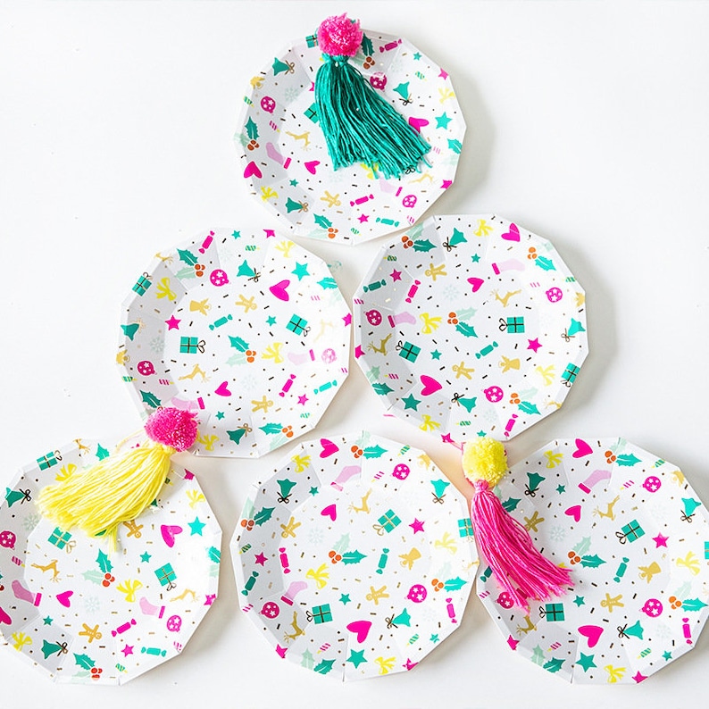 May include: Six white paper plates with a festive holiday pattern of hearts, gifts, and holly. Each plate is adorned with a colorful tassel and pom-pom in shades of pink, yellow, and teal. Perfect for a holiday gathering.