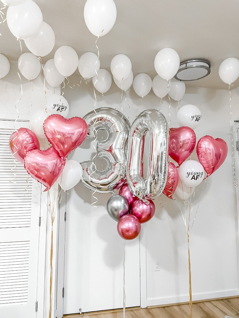 Może przedstawiać: Srebrne balony z numerem 30, r&oacute;żowe balony w kształcie serca i białe balony z napisem "young AF".