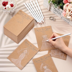 May include: A stack of brown kraft paper cards with the text Guess the Dress and a white line drawing of a wedding gown. Several white pencils are included, and one is being used to draw on a card. The cards feature various dress styles.