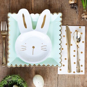 May include: Easter-themed table setting with a bunny-shaped plate, mint green square plate, and gold cutlery. A white napkin with gold polka dots and a small nest with an egg complete the festive arrangement on a wooden surface.