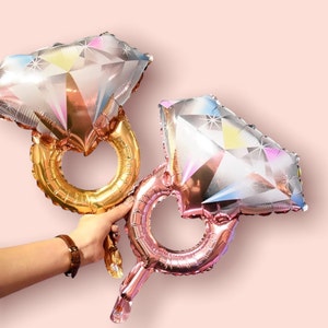 May include: Two foil balloons shaped like diamond rings. The balloons are iridescent and have a pink and gold colour scheme.