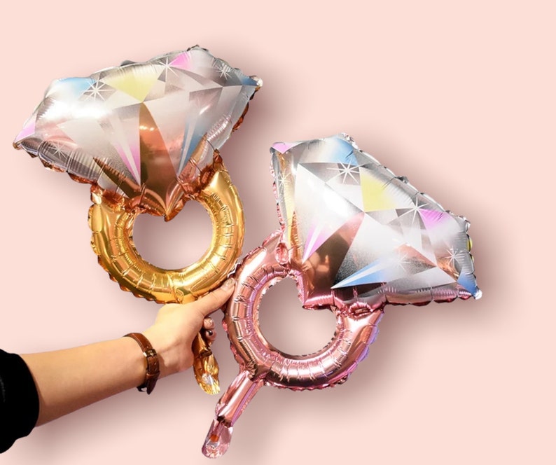 May include: Two foil balloons shaped like diamond rings. The balloons are iridescent and have a pink and gold colour scheme.