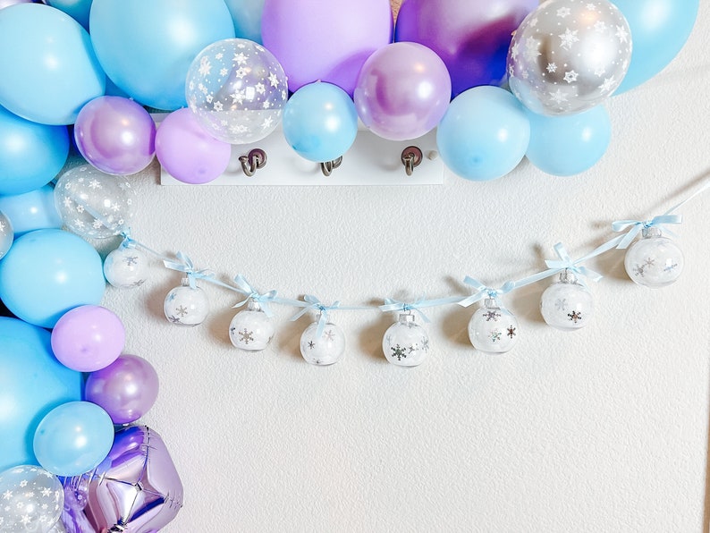 May include: A festive balloon arch in shades of light blue, lavender, and silver, with clear balloons featuring white snowflake patterns. A garland of small, clear ornaments with snowflake designs hangs below.