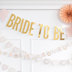 K&ouml;nnte beinhalten: Goldene Folien-Girlande "Bride to Be" mit rosa-wei&szlig;en gestreiften Flaggen und einer Girlande aus wei&szlig;en Herzen mit goldenen Akzenten und rosafarbenen Kuchenmotiven.