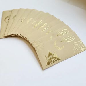 May include: A stack of tan-colored greeting cards with gold foil accents. The cards feature floral and mosque designs, along with Arabic script. The cards are arranged in a fan shape, showcasing the intricate details.