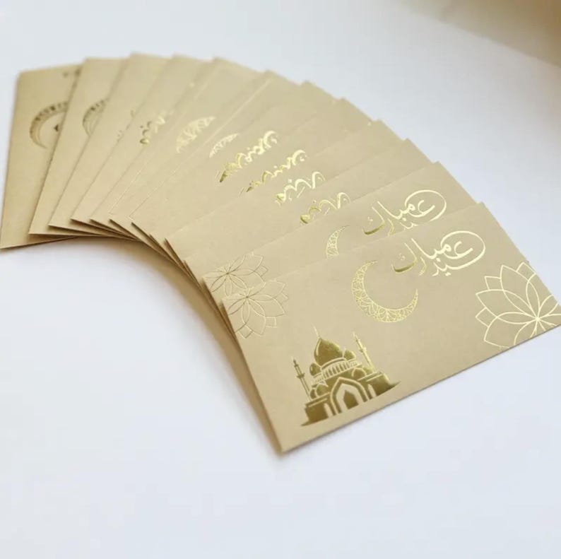 May include: A stack of tan-colored greeting cards with gold foil accents. The cards feature floral and mosque designs, along with Arabic script. The cards are arranged in a fan shape, showcasing the intricate details.