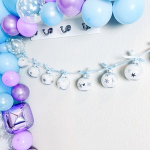 May include: A festive display featuring a balloon arch in shades of blue, purple, and silver. A garland of clear ornaments with snowflake designs hangs below. The balloons are arranged near a white wall with a row of hooks.