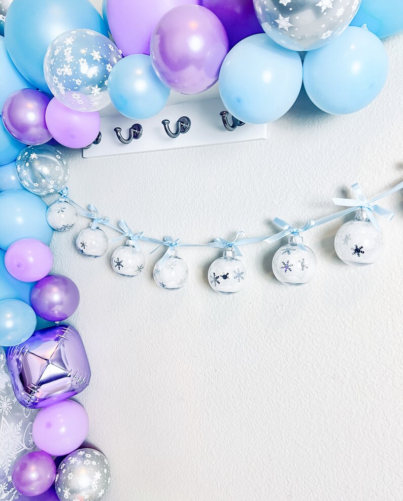 May include: A festive display featuring a balloon arch in shades of blue, purple, and silver. A garland of clear ornaments with snowflake designs hangs below. The balloons are arranged near a white wall with a row of hooks.